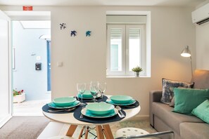 Un coin repas avec une table ronde dressée pour quatre personnes, un canapé avec des coussins verts, une fenêtre avec des stores et une petite plante posée sur le rebord de la fenêtre.