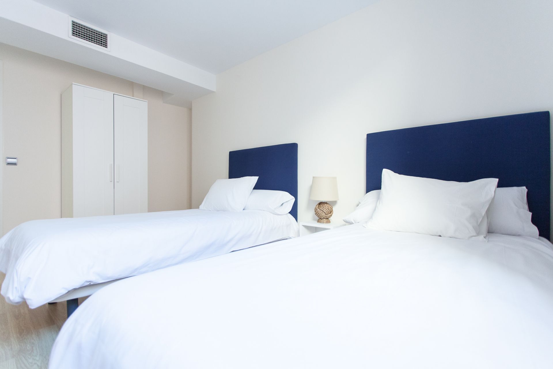 Una habitación de hotel con dos camas individuales, ropa de cama blanca, cabecero azul, una lámpara de noche y una puerta con manija.