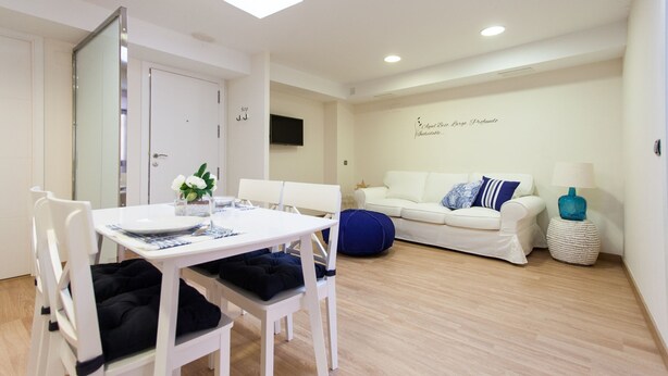 A modern, well-lit room with a dining table, white chairs, a sofa, and a small side table with a lamp. - Albareda 6 - Two Bedroom Apartment, Sleeps 4 (Sevilla)