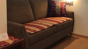 A brown sofa with a patterned throw blanket and multiple pillows. A small wooden table with a sign is visible to the left. - Affittacamere Mulino Square 26 (Matera)