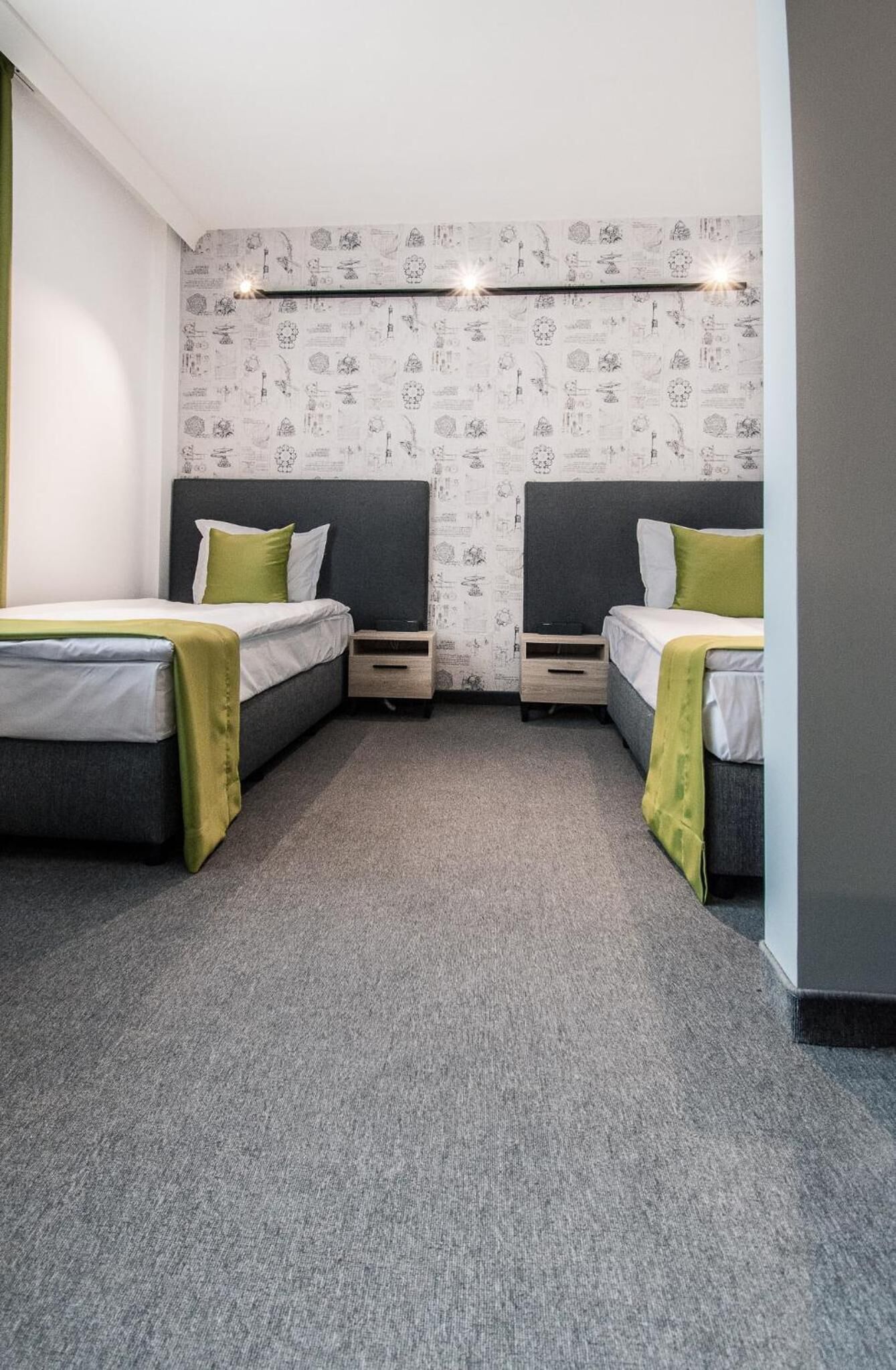 Une chambre avec deux lits, chacun doté d’une tête de lit sombre et d’un oreiller vert. Les murs sont recouverts d’un papier peint à motifs, et il y a deux tables de chevet avec des tiroirs. Le sol est recouvert d’une moquette.