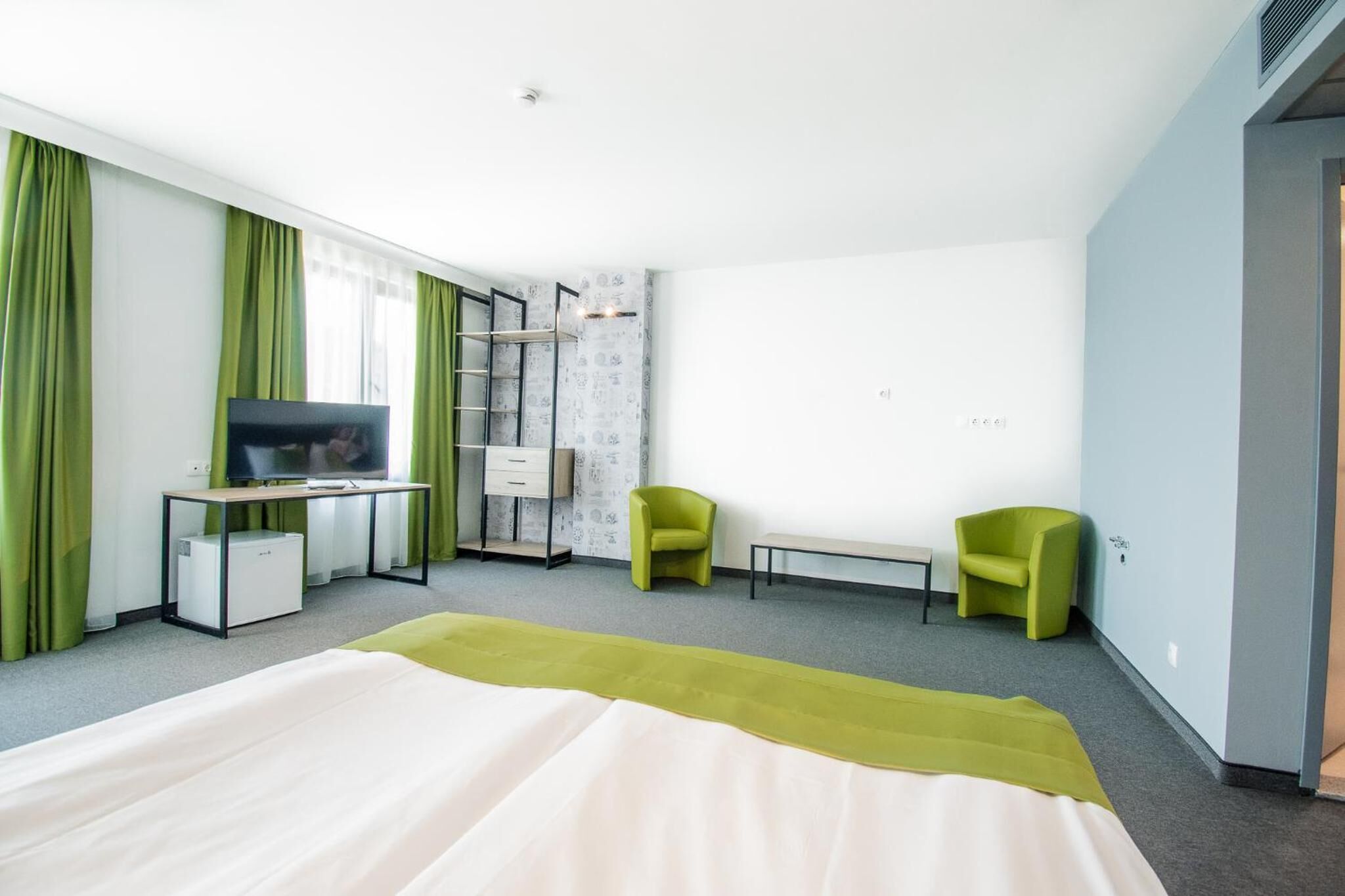A hotel room with a bed, a desk with a TV, two green chairs, a small table, and a wardrobe.