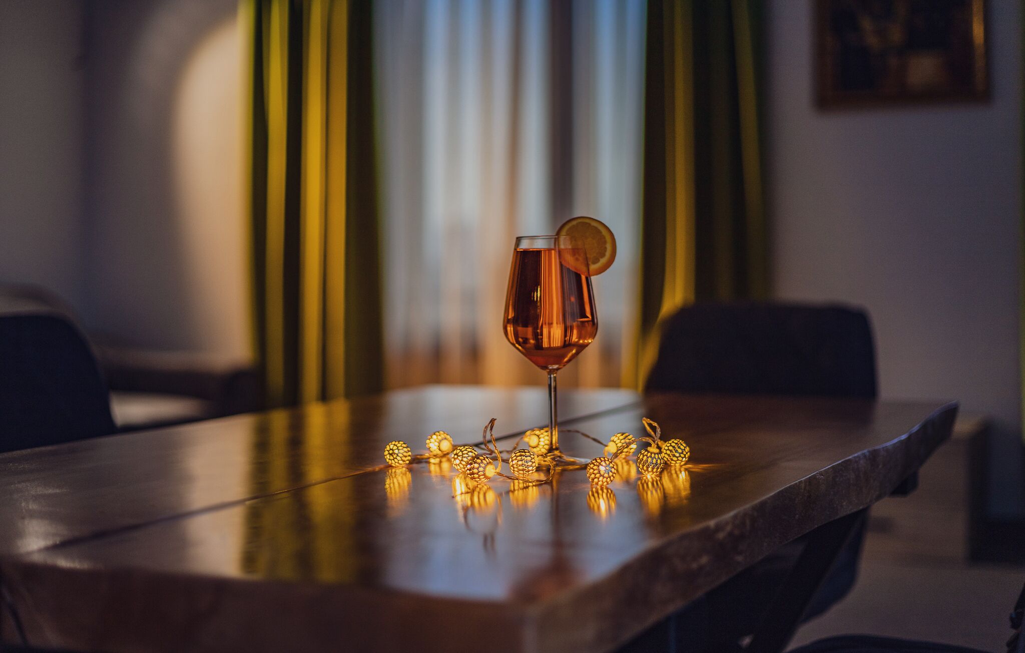 A table with a glass of amber-colored liquid, possibly a cocktail, garnished with a slice of orange. The table is surrounded by chairs, and there are string lights with spherical bulbs on the table's surface.
