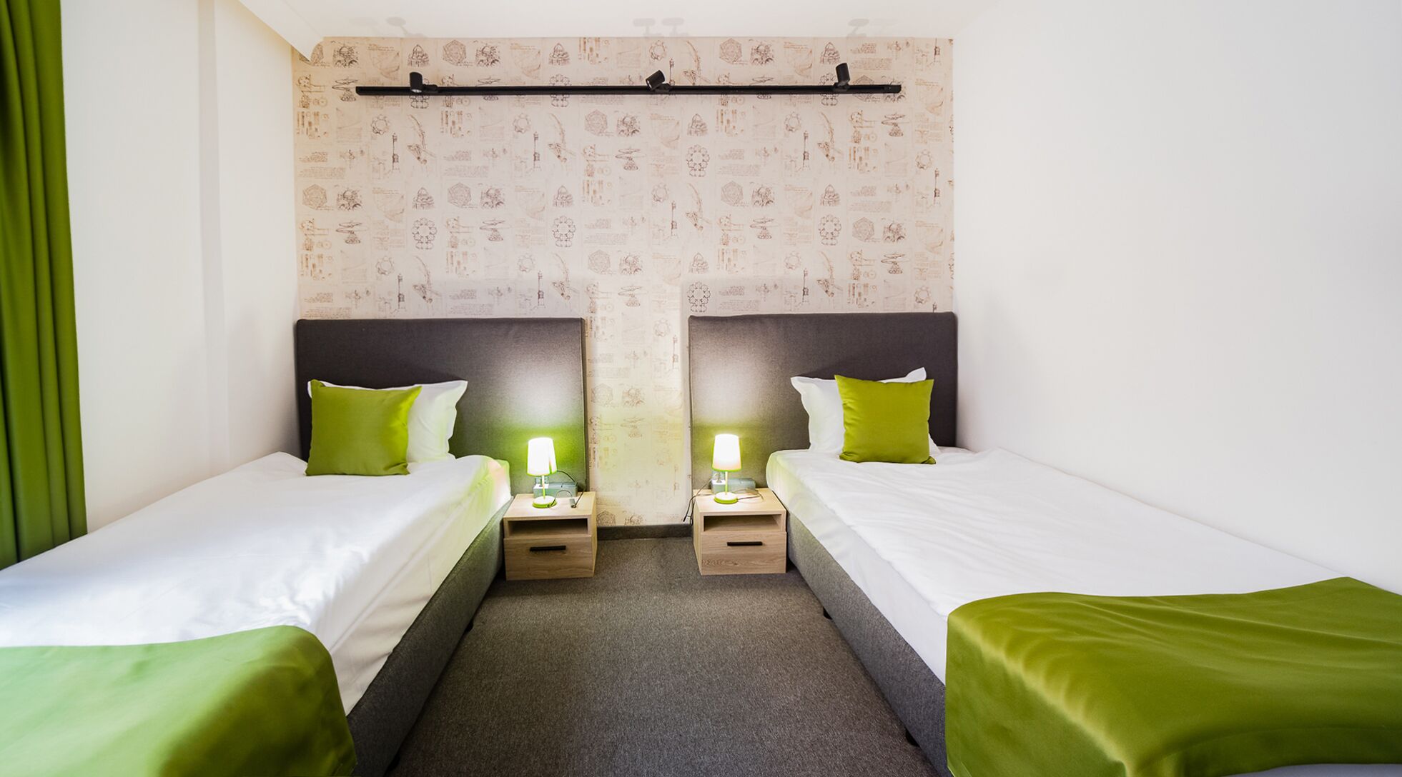A twin room with two beds, each with a green pillow, a bedside table with a lamp, and a patterned wallpaper.