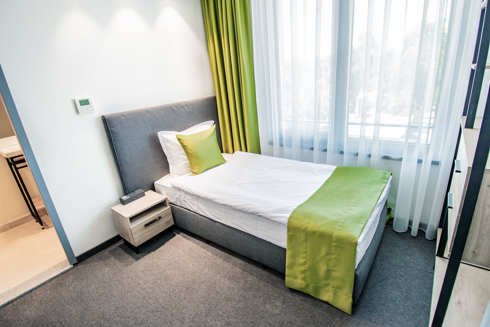 Una habitación de hotel con cama, mesita de noche y ventana con cortinas translúcidas.