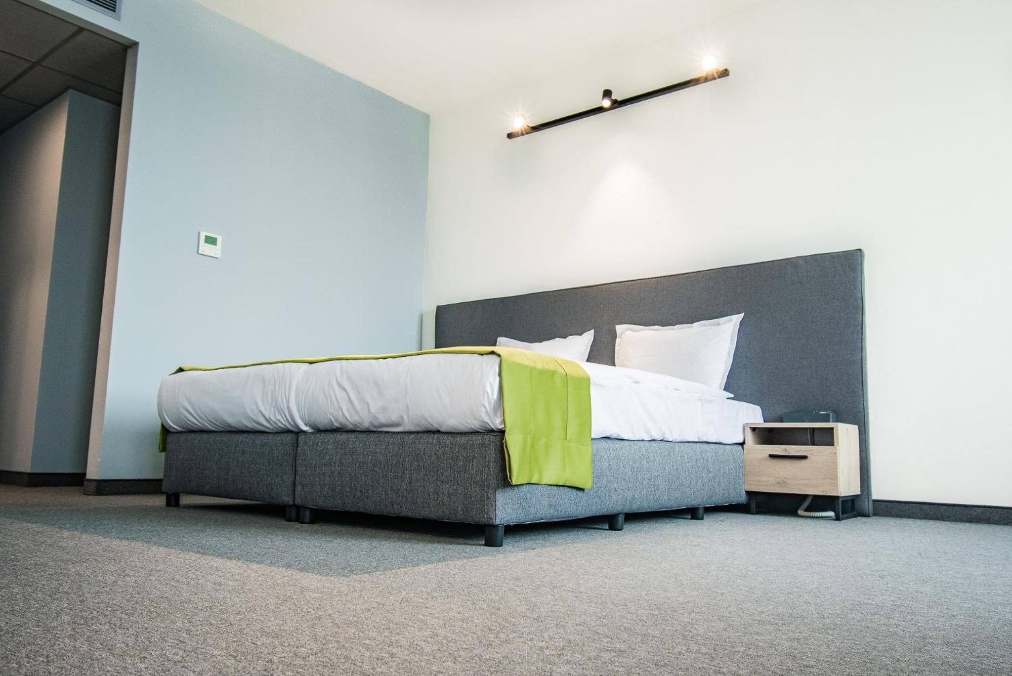 A modern hotel room with a grey bed, white bedding, a green throw, a bedside table with a lamp, and a wall-mounted light fixture.
