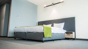 A modern hotel room with a grey bed, white bedding, a green throw, a bedside table with a lamp, and a wall-mounted light fixture.