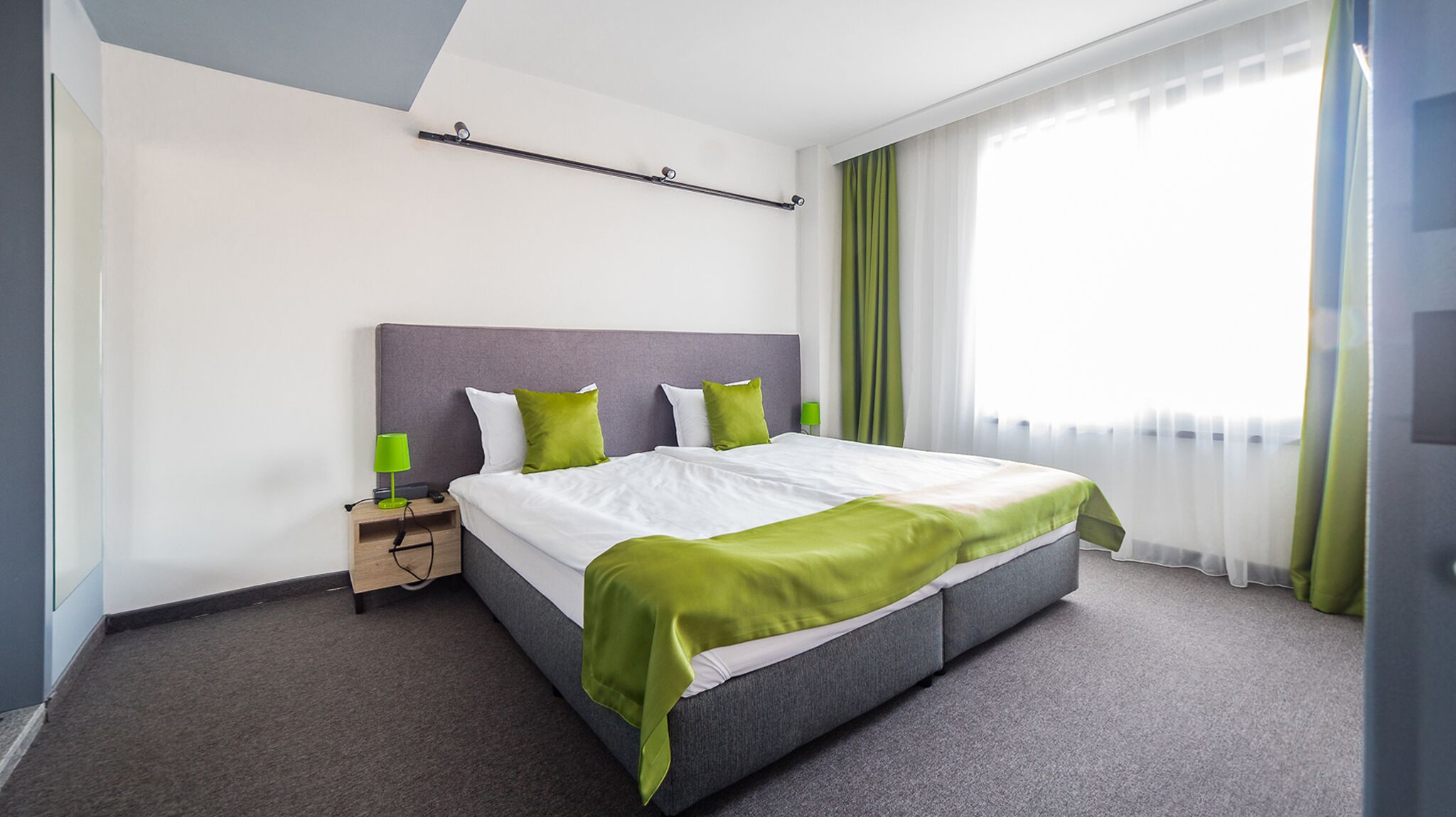 A modern hotel room with a large bed, a nightstand with a lamp, a window with sheer curtains, and a headboard.