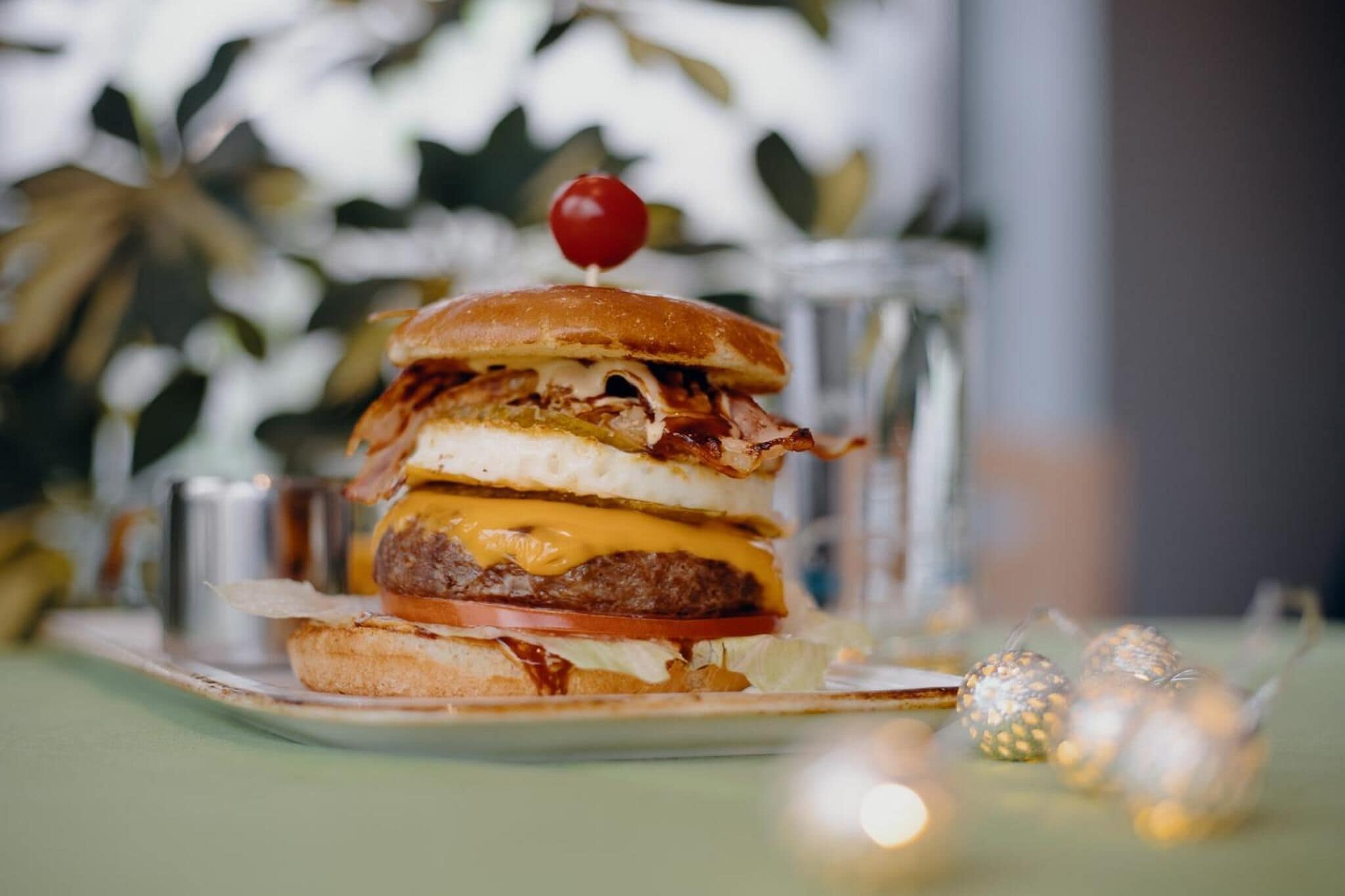 Un sándwich con cereza encima, sobre un plato con un vaso de agua y luces decorativas al fondo.