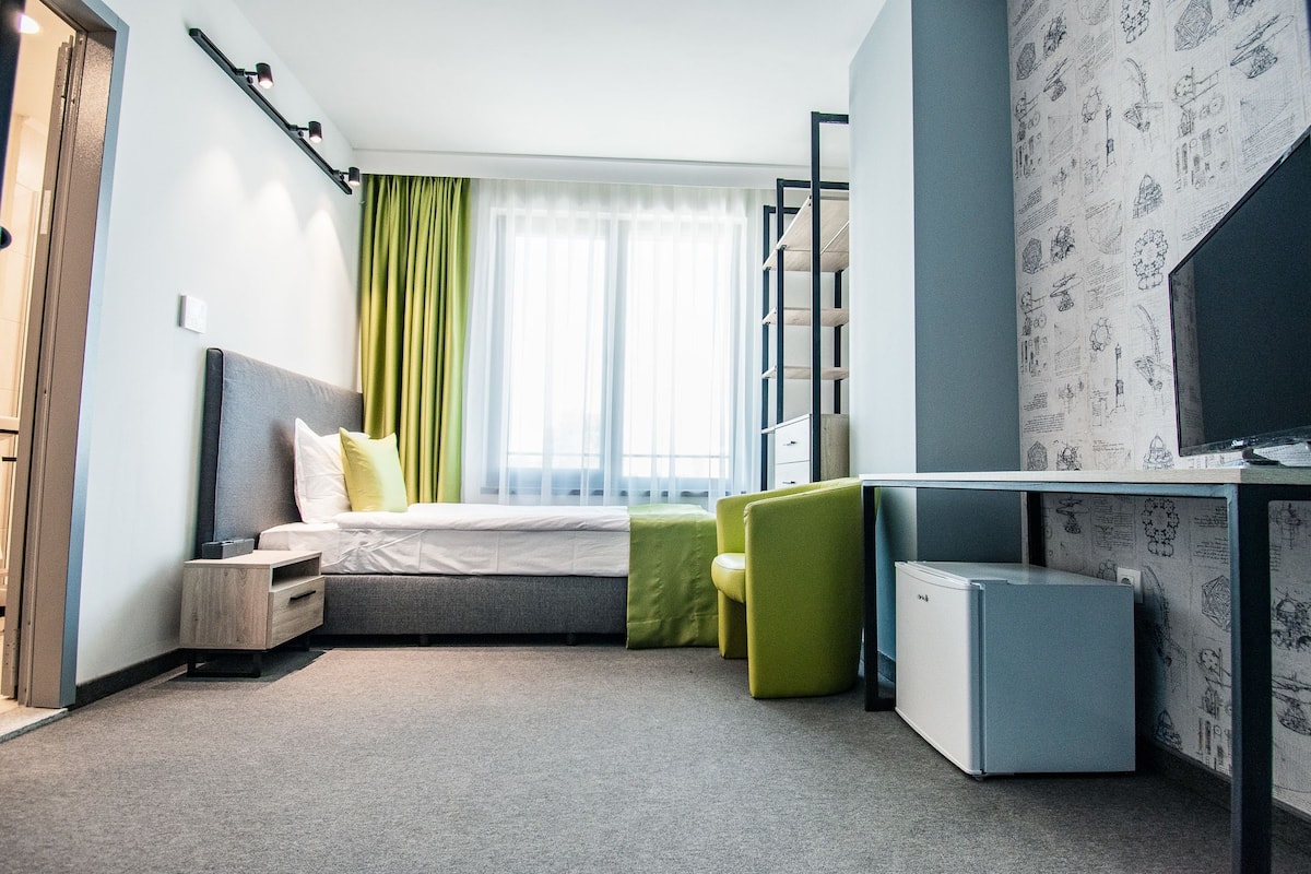 Une chambre d’hôtel moderne équipée d’un lit, d’un bureau, d’un petit réfrigérateur et d’une fenêtre avec des rideaux.