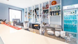 A bar area with a long counter, shelves stocked with various bottles, a coffee machine, and a refrigerator filled with water bottles. - A&M Hotel - Plovdiv (Plovdiv)