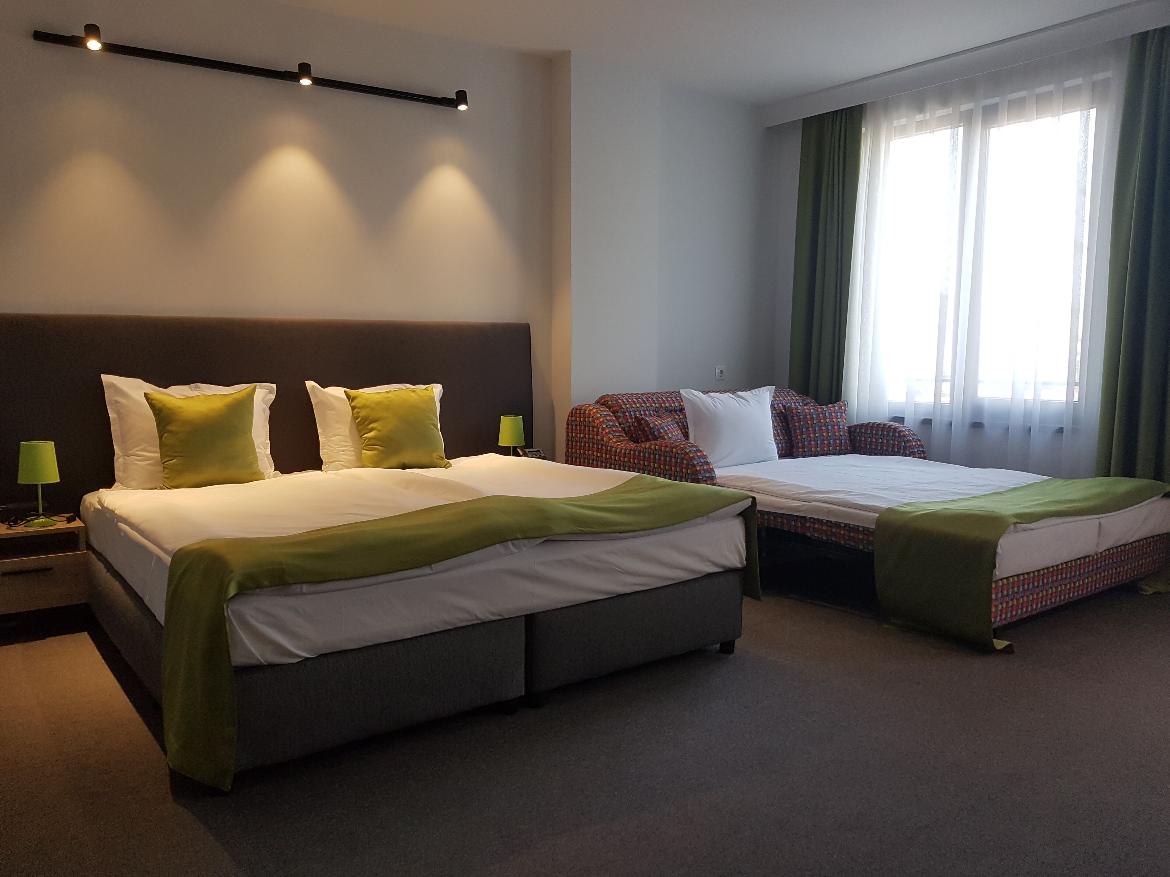 A hotel room with two beds, a headboard, a bedside table with a lamp, and a window with curtains.