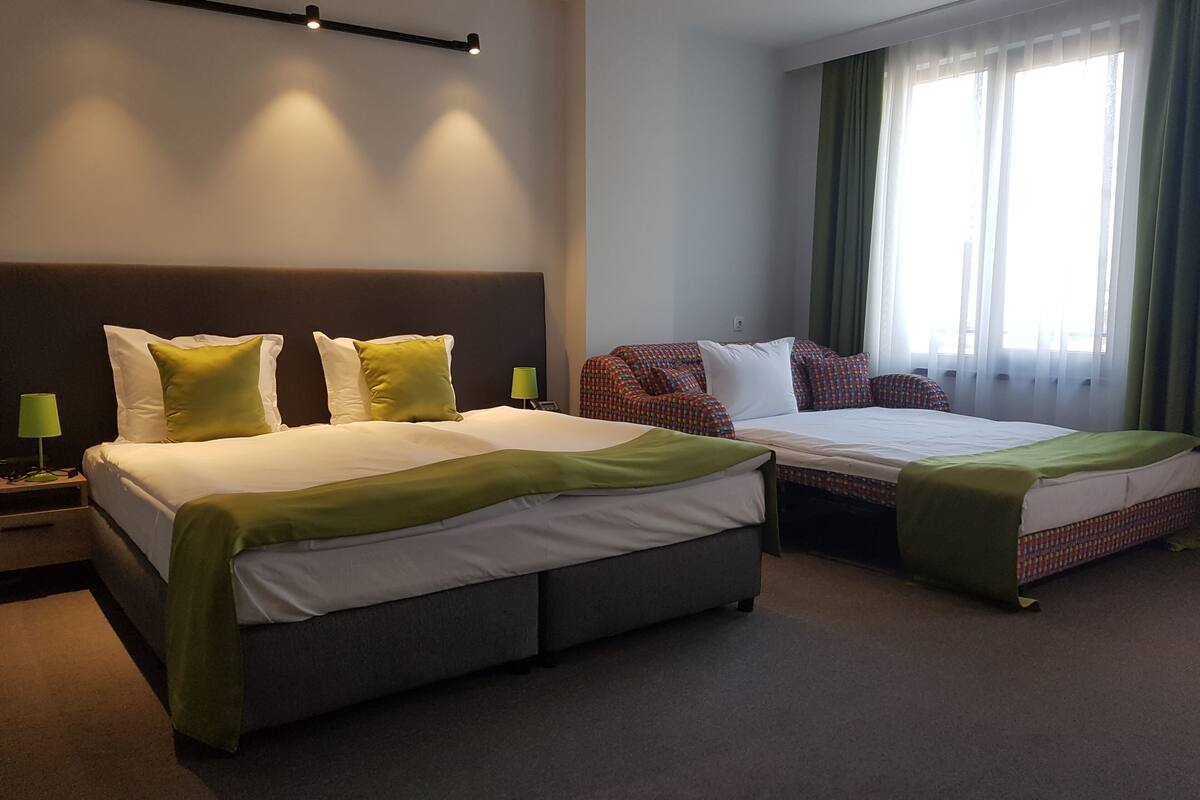 A hotel room with two beds, a headboard, a bedside table with a lamp, and a window with curtains.