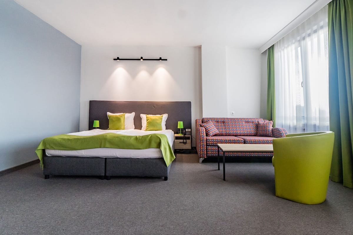 Une chambre d’hôtel avec un lit, un canapé, une chaise et une grande fenêtre avec des rideaux.
