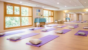 A yoga studio with multiple mats and pillows arranged on a wooden floor, large windows, and a table with a blue tablecloth.