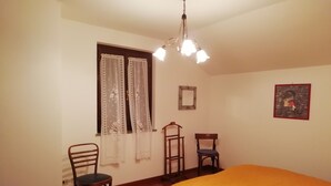 A bedroom with a bed, two chairs, a wooden dresser, a window with curtains, a mirror, and a chandelier. - La via del bosco Apartment in a villa (Casteldelfino)