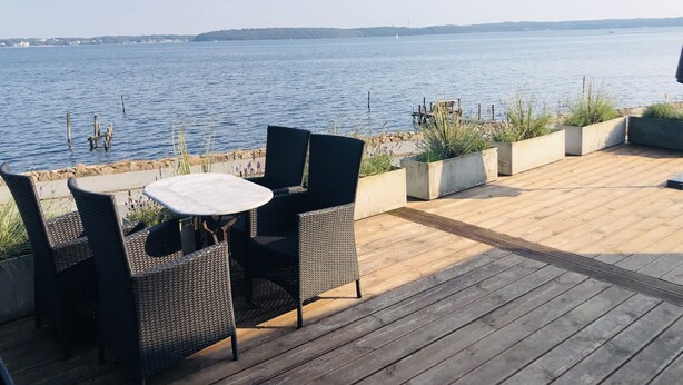 A wooden deck with wicker furniture and planters, overlooking a calm body of water. - Villa Alouisia (Krusaa)