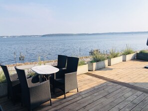 Una terraza de madera con muebles de mimbre y macetas, con vistas a una masa de agua tranquila.