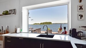 A kitchen with a large window overlooking a body of water, a sink, and a coffee maker. - Villa Alouisia (Krusaa)