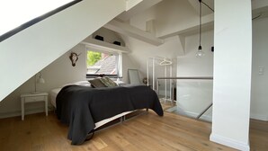 A modern bedroom with a sloped ceiling, a bed with a dark bedspread, a bedside table with a lamp, a window with a view of greenery, and a glass railing in the corner. - Villa Alouisia (Krusaa)