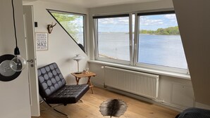 A room with a view of a lake, a black leather chair, a small wooden table, a lamp, and a fur rug on the floor. - Villa Alouisia (Krusaa)