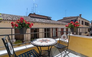 Ein Balkon mit rundem Tisch und Stühlen, Blumentöpfen mit roten Blüten und einem Ziegeldach im Hintergrund.