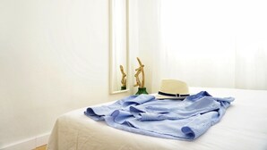 Een net opgemaakt bed met een wit laken, een blauw gestreept overhemd gevouwen en een beige hoed op het bed. Er hangt een lijst met een foto aan de muur en op een klein tafeltje naast het bed staan twee decoratieve houten beeldjes.