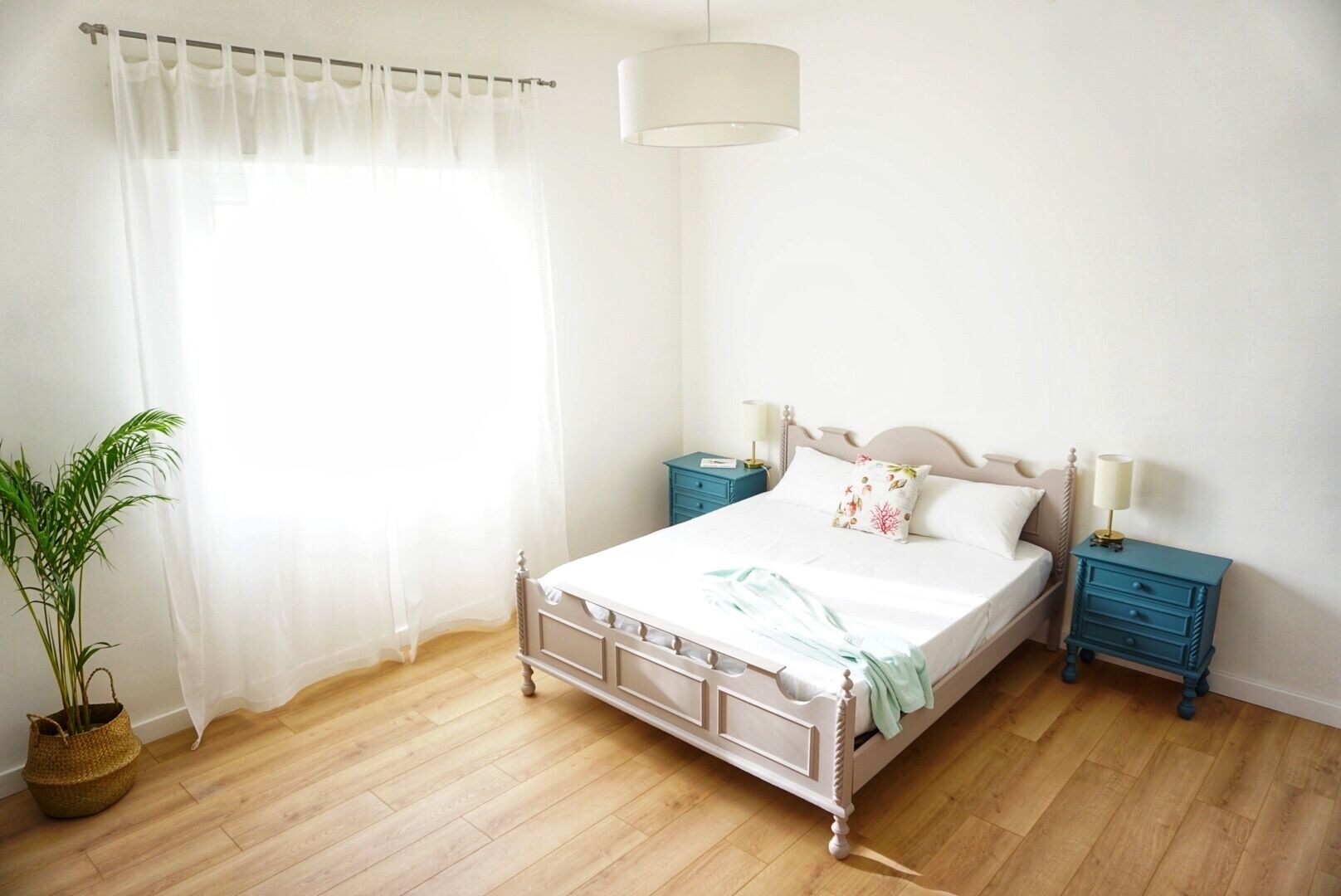A bedroom with a bed, two nightstands, a potted plant, and a window with sheer curtains.