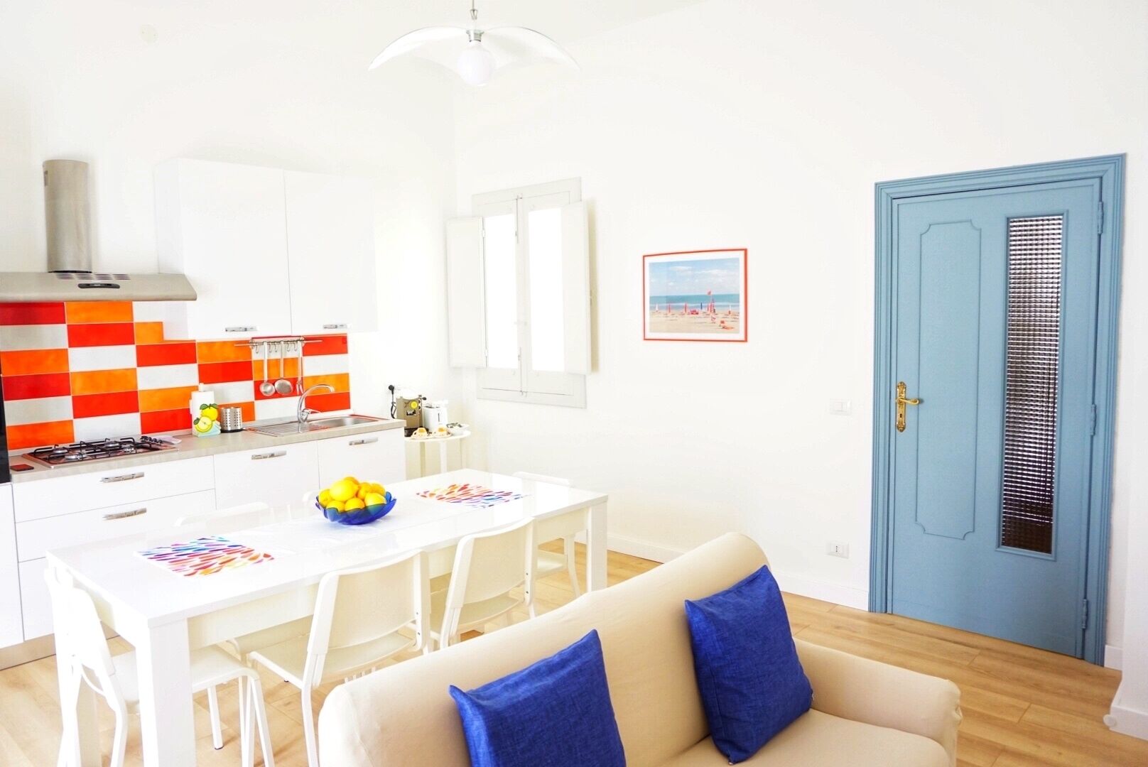 A modern kitchen with white cabinets, a gas stove, and a colorful backsplash. There is a dining table with chairs and a blue door with a window.