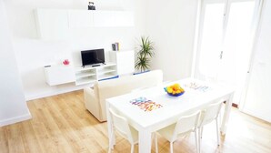 Een moderne, minimalistische eetruimte met een witte tafel en stoelen, een fruitschaal en een flatscreen televisie op een wit meubel.