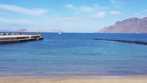 A coastal scene with a pier extending into calm blue waters, a sandy beach, and distant mountains under a clear sky. - Saltpeter: 1st Beach Line (Caleta de Famara)