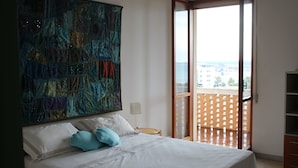 Un dormitorio con cama, almohadas, un cuadro abstracto grande, una mesita de noche y vistas a un balcón con valla de madera.