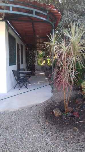 Uma área de estar ao ar livre coberta, com mesa e cadeiras, cercada por cascalho e plantas.