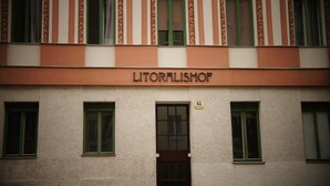 Ein Gebäude mit dem Schild "LITORALISCHOF" über dem Eingang. Im Erdgeschoss befinden sich drei Fenster, jedes mit einem grünen Rahmen. Ein gelbes Schild mit der Nummer "65" ist auf der rechten Seite des Gebäudes sichtbar.