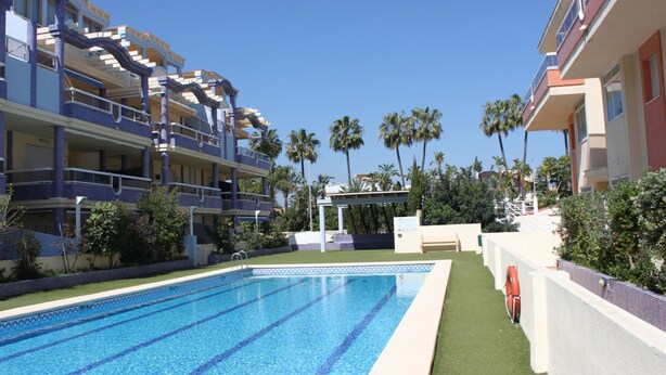 En swimmingpool med krystalklart, blå vand, omgivet af en hvid poolterrasse og et område med grøn kunstgræs.