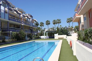 Une piscine à l’eau d’un bleu cristallin, entourée d’une terrasse blanche et d’une zone recouverte de gazon synthétique vert.