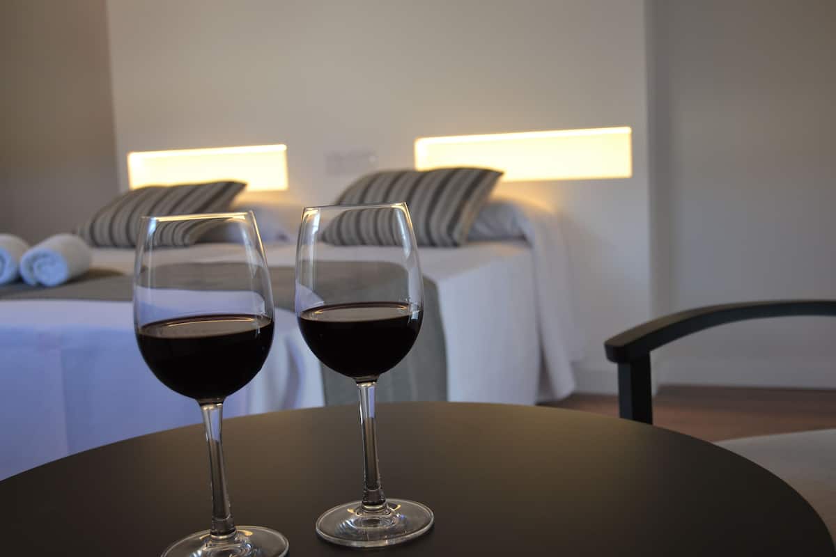 Two glasses of red wine on a table in a hotel room with a bed and a nightstand in the background.