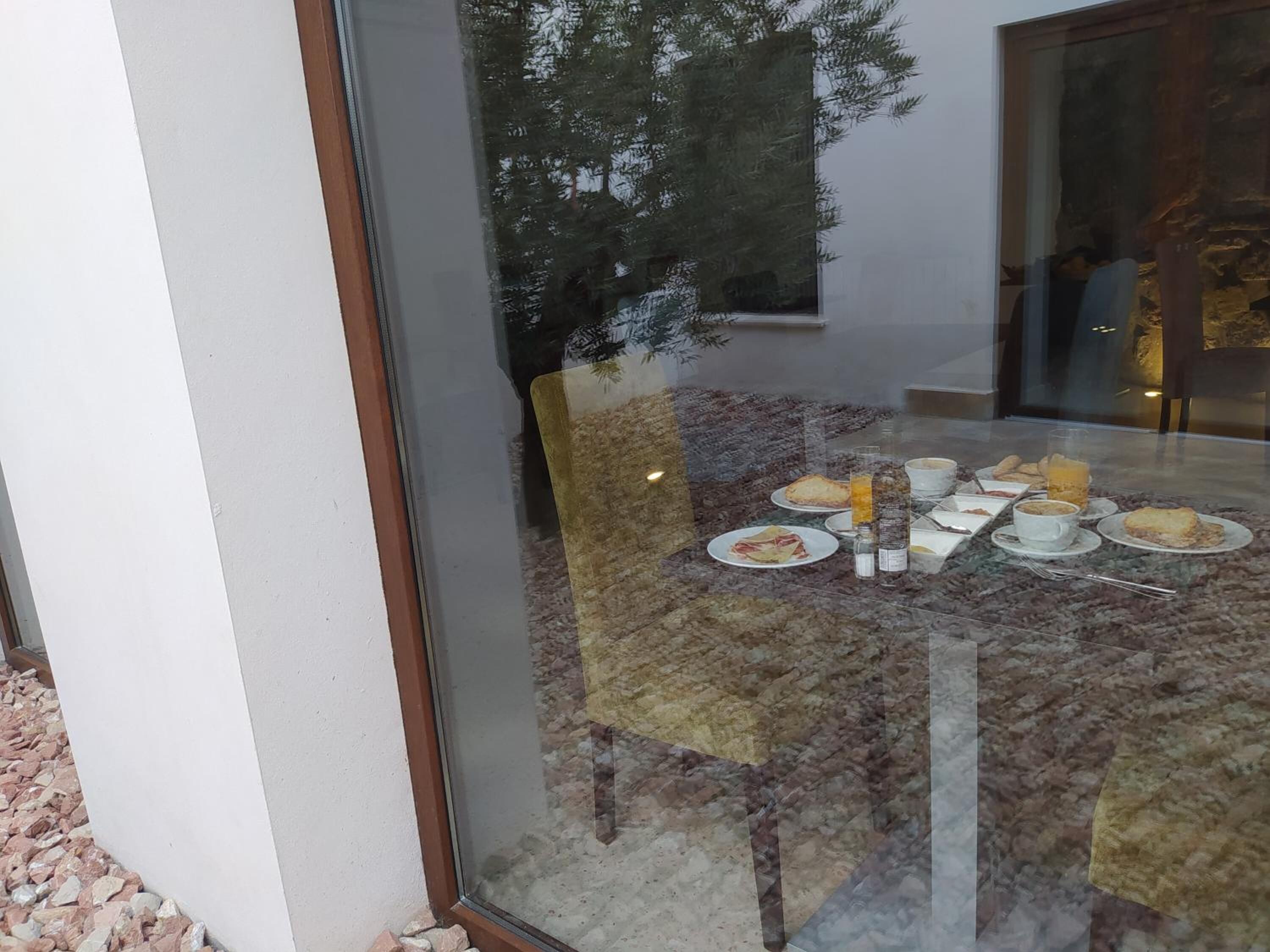 Un comedor con una mesa y sillas de vidrio, diversas opciones para el desayuno y vista a árboles afuera.