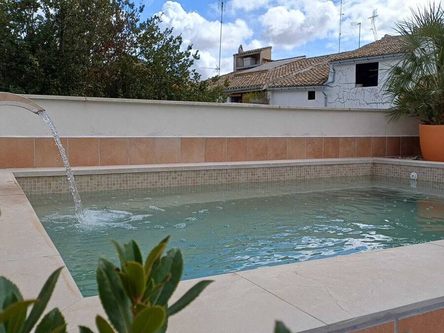Una piscina rectangular al aire libre con borde de azulejos, un chorro de agua y una planta en maceta a la derecha.