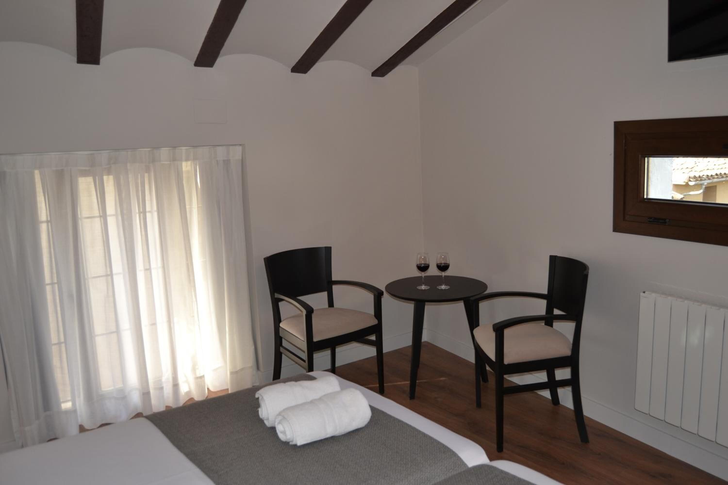 A hotel room with a bed, two chairs, a small table with wine glasses, and a window with curtains.