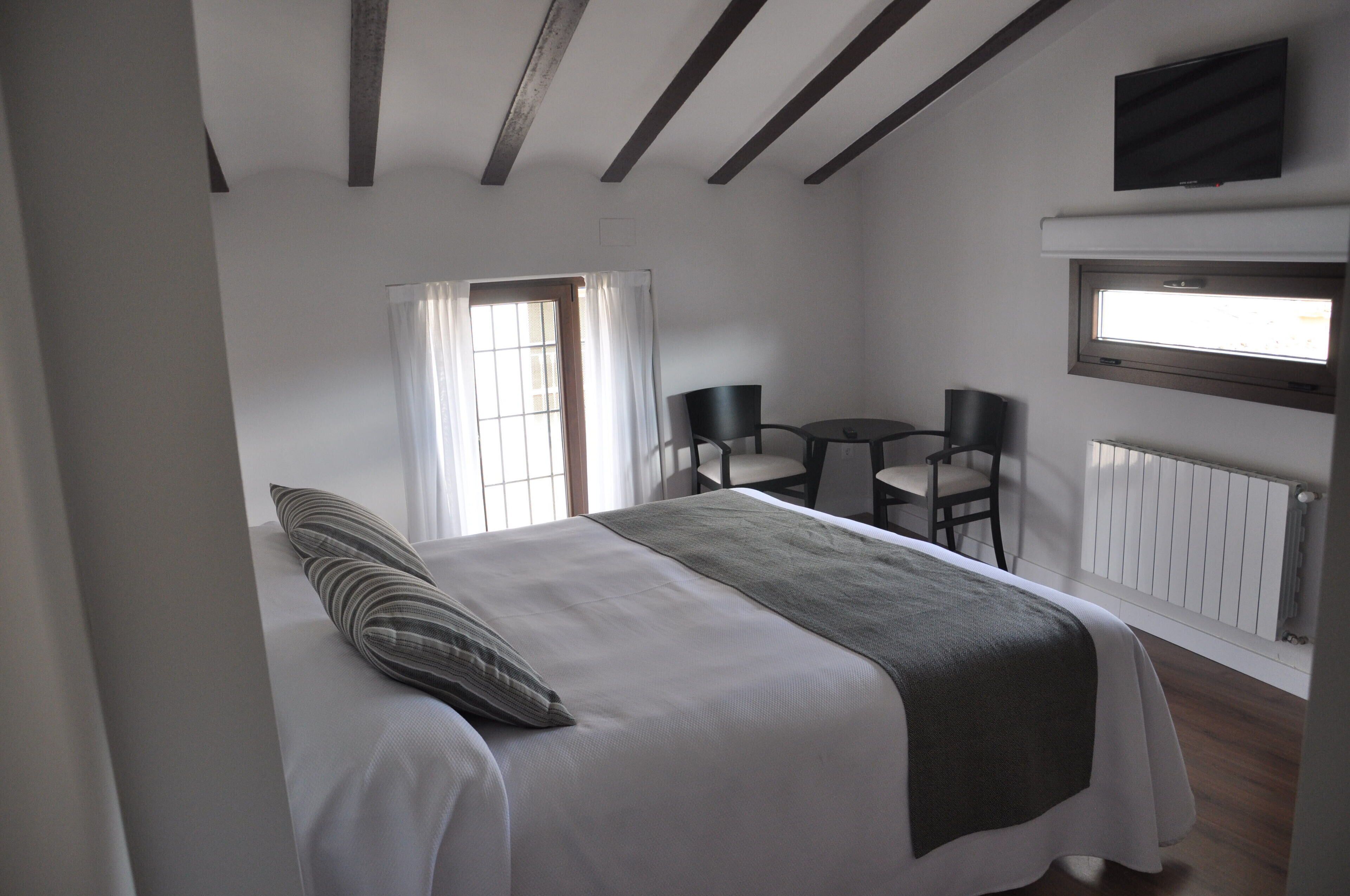 Un dormitorio con una cama, una ventana, una mesa con sillas y un televisor montado en la pared.