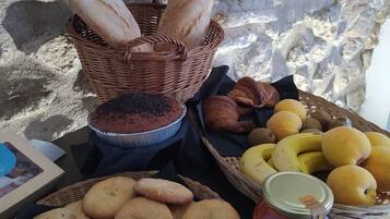 Una mesa con varios productos horneados, incluyendo pan, cruasanes, galletas y un pastel, junto con una cesta de fruta y un frasco de mermelada.