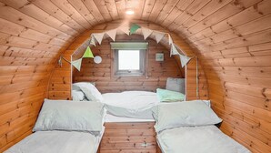 A small, cozy attic bedroom with wooden walls, a window, two beds, and a ceiling fan. - Rivendell Glamping Pod - UK11881 (Holsworthy, near Launceston)