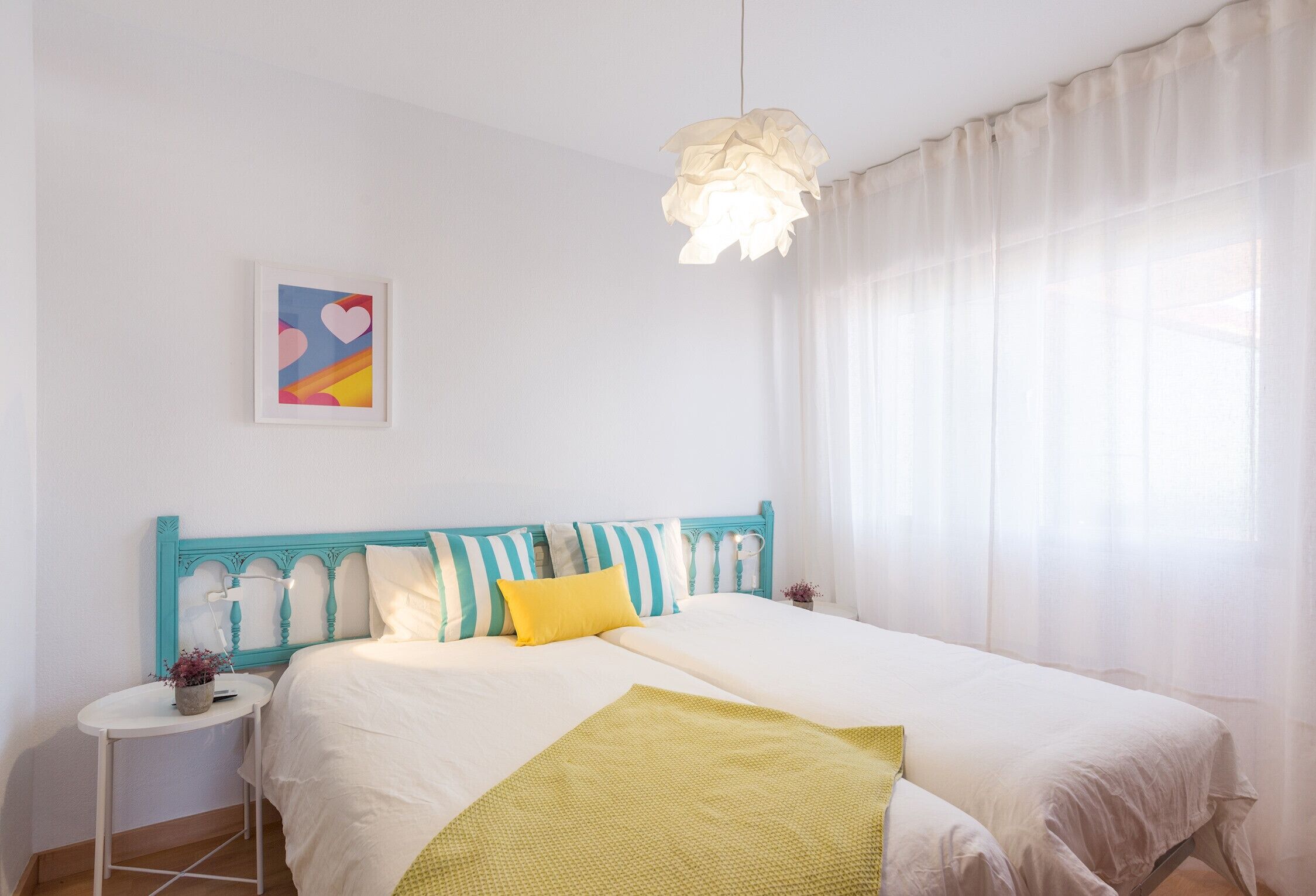 A bedroom with a bed, a bedside table, a hanging light fixture, and a window with curtains.