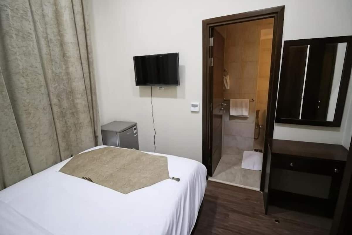 Quarto de hotel com cama, televisão, escrivaninha, espelho e chuveiro.