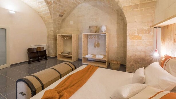 A cozy room with a stone vaulted ceiling, a bed with a brown blanket, a wooden bench, and a shelf with towels and a basket. - Balbo 7 Apartment - Lecce Selection (Lecce)