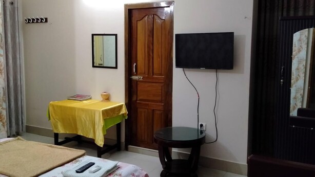 A room with a bed, a small table with a yellow tablecloth, a chair, a TV mounted on the wall, and a wooden door. - Hotel Charu (Barisal)