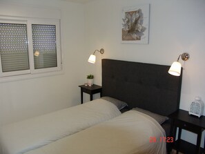 Ein Schlafzimmer mit einem Bett, zwei Nachttischen, einer Wandlampe, einem Fenster mit Jalousien und einem gerahmten Kunstwerk.
