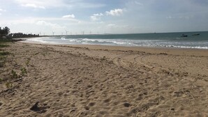 A sandy beach with footprints leading to the water, a clear sky, and wind turbines in the distance. - Casa das Rendas Exclusive Bungalows (Amontada)