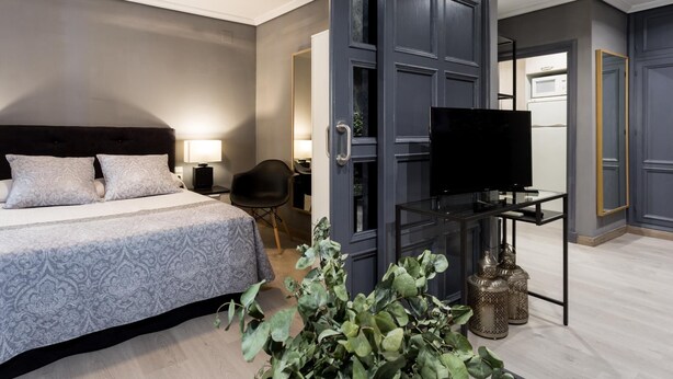 A modern hotel room with a large bed, a TV, a desk, and a mirror. - Holi-Rent Gran Poder (Sevilla)